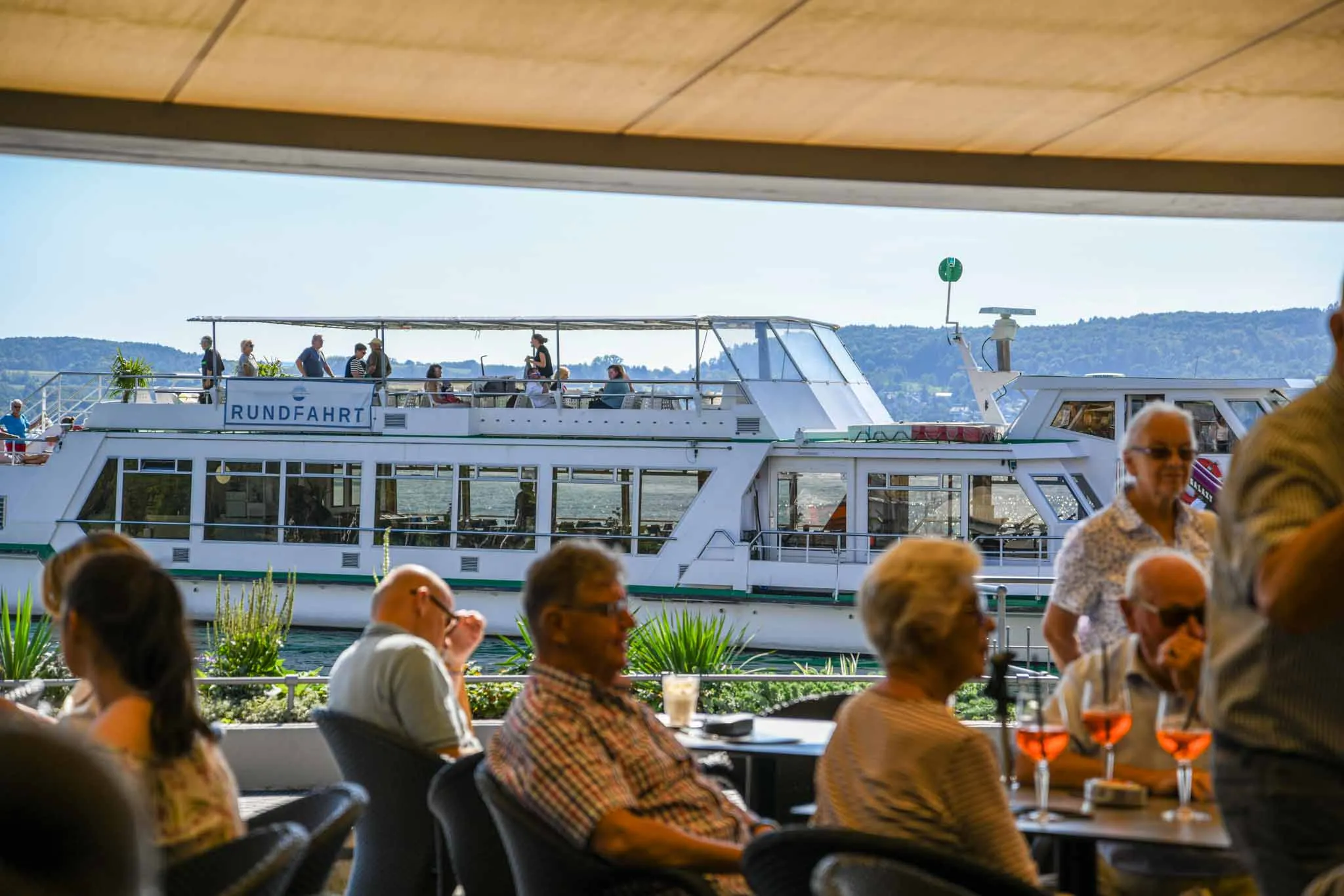 Gäste auf der Terrasse, dahinter legt ein Rundfahrtschiff an der Promenade an