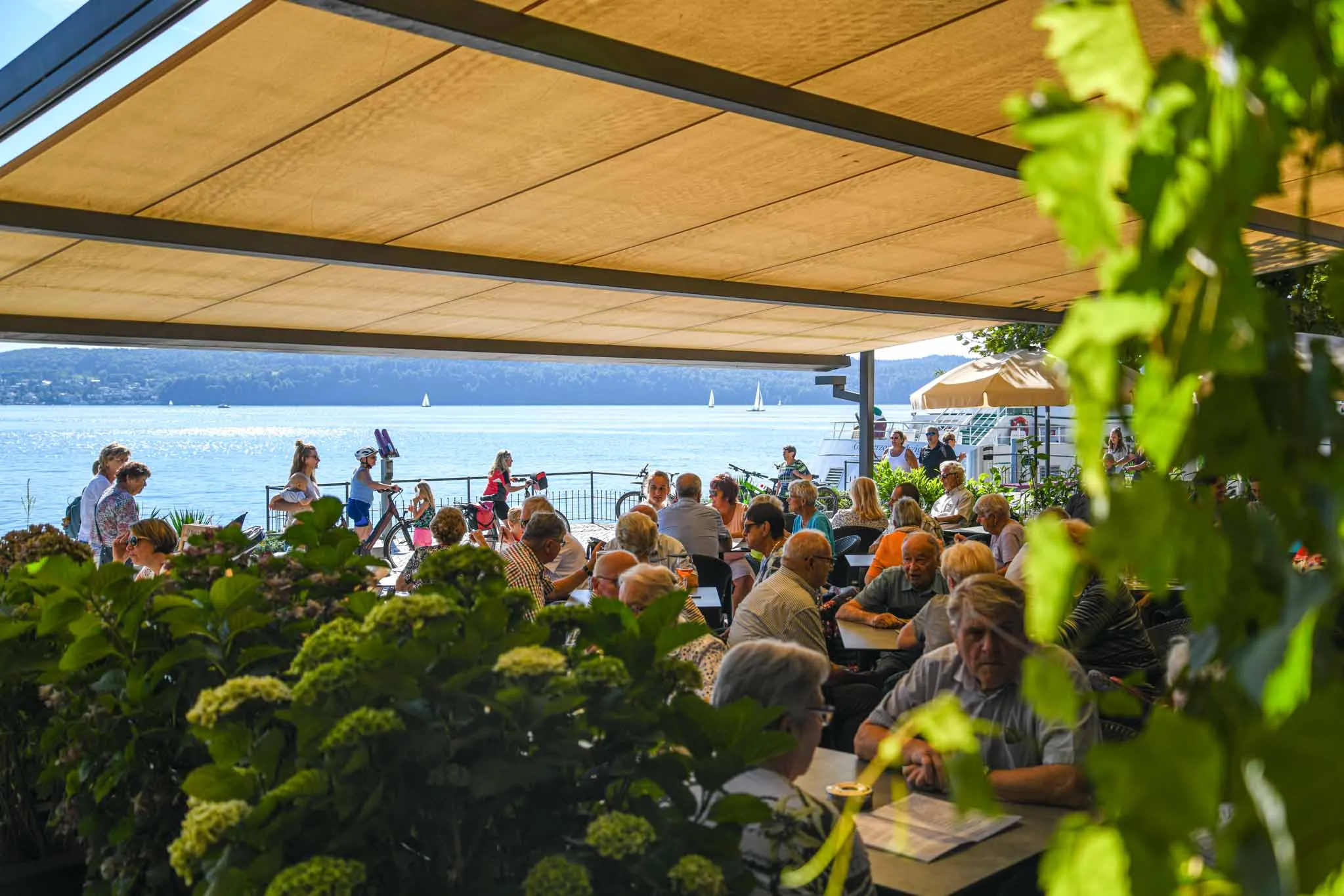 Die voll besetzte Terrasse des Café Walker unter der Markise, dahinter der Überlinger See mit Segelbooten